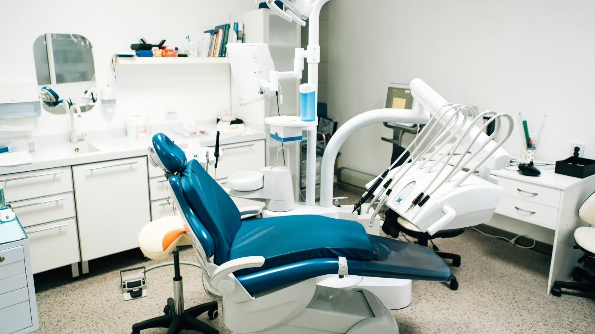 The image shows a dental office interior with a large dental chair and equipment, featuring a clean, professional environment with white walls and cabinets.